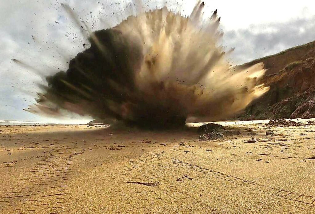 An explosion has sent sand blasting in all directions in the top of the image. The foreground is a sandy beach.