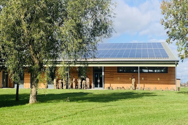 A rectangle light wooden building with solar panels across the roof. The door is black and there are windows at the right-hand side of the building. There are nine troops facing the building infront and a tree infront of the building with grass.