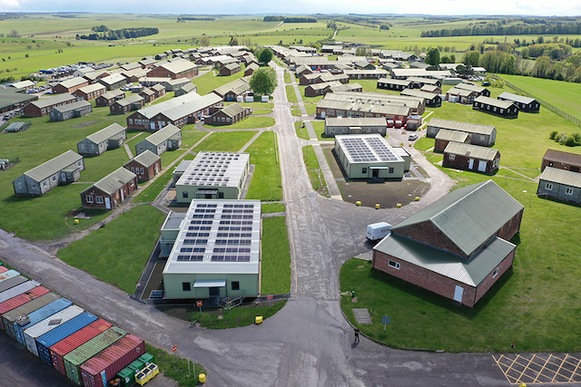 Aeriel view of the three new Net-Zero Carbon efficient buildings at Westdown Camp. The buildings are grey rectangle blocks with a flat roof and solar panels at the top. There is grass around them and a road. There are several other office blocks in brown around the buildings.