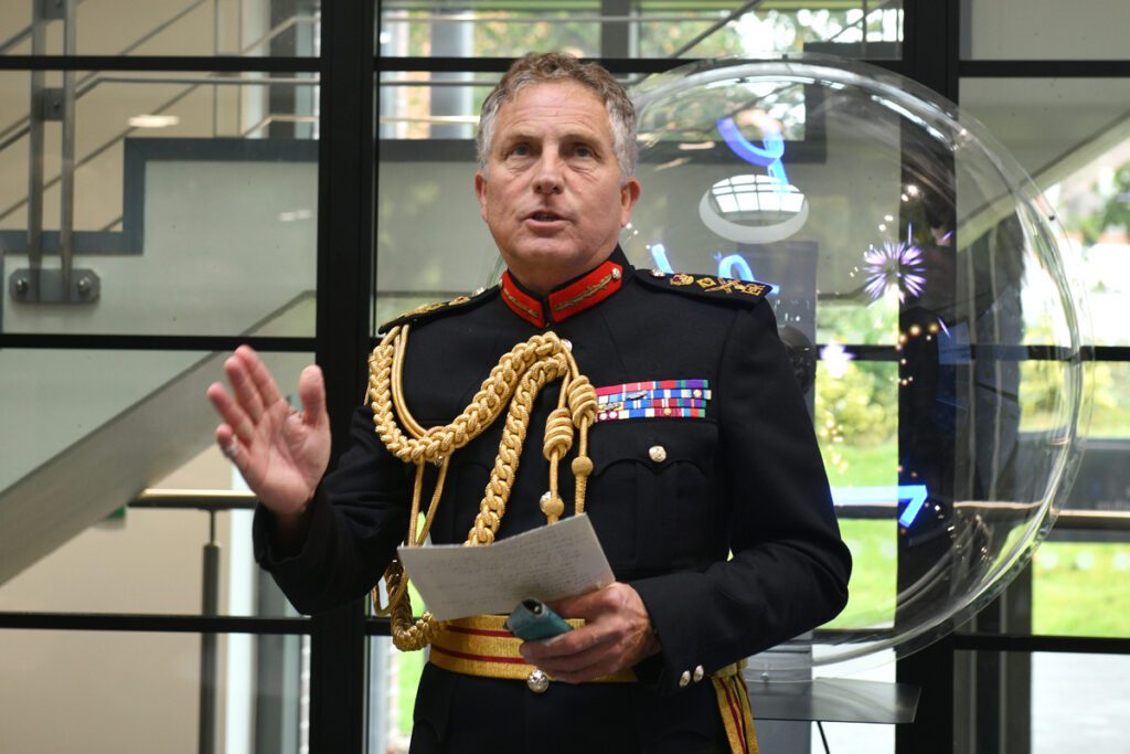 General Sir Nick Carter, a white man wearing a black military uniform with gold braid, stands in front of a glass wall. He is holding a sheet of paper as he makes a speech.