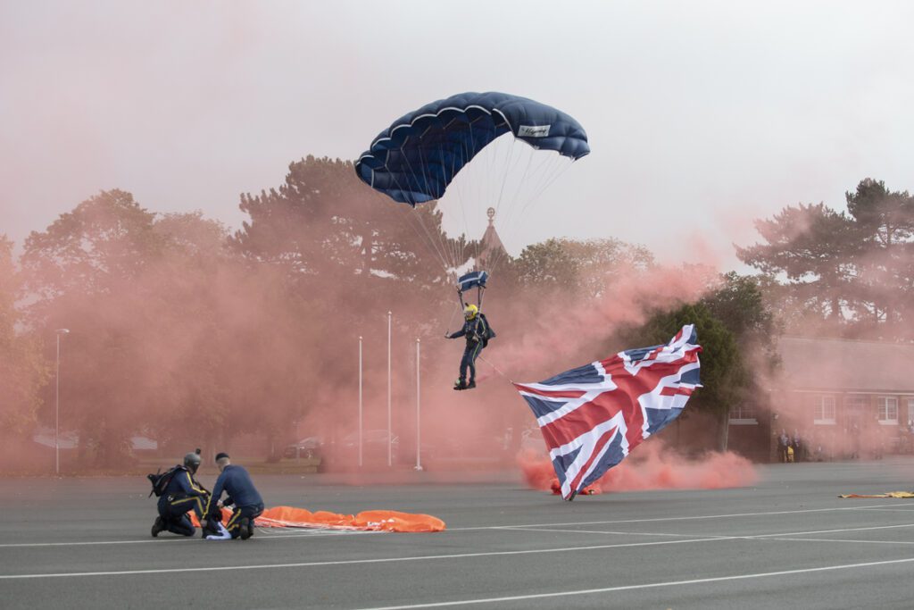 A parachutist comes in to land on a large open concrete area with buildings behind. He has a dark parachute and a union flag flies from him. Another parachutist has already landed nearby.