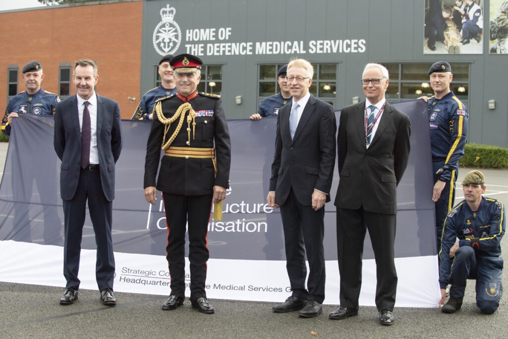 Three men in suits stand around another man in black military uniform with gold braid and a hat. They stand in front of a large flag with the DIO logo on it, which is being held by a number of men in parachuting suits. Behind them is a large building with text on the side reading "Home of the Defence Medical Services".