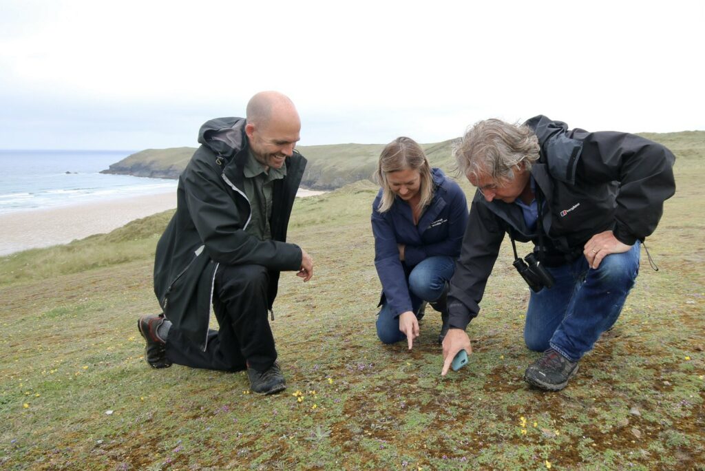 Three people in dark clothing kneel on the ground looking at something between them. They are on a grassy area with the sea behind them.