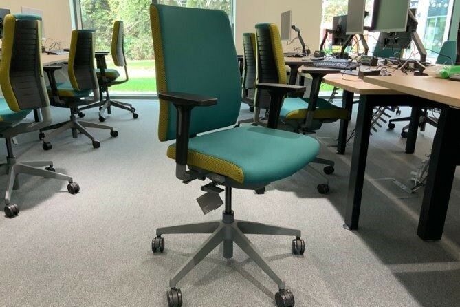 The photo shows an office space with a chair in the centre of the image. The chair has green cushioning, adjustable levers and wheels. Either side of the chair are desks with equipment including keyboards and monitors on them.