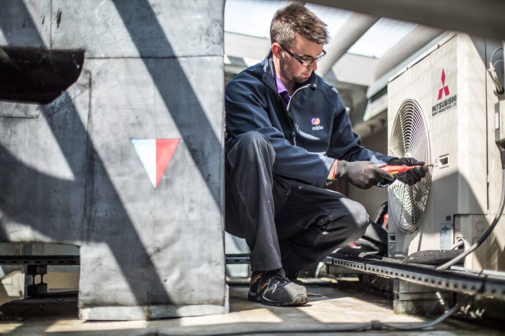 A Mitie engineer kneels next to what may be an air conditioning unit. He seems to be opening the cover.