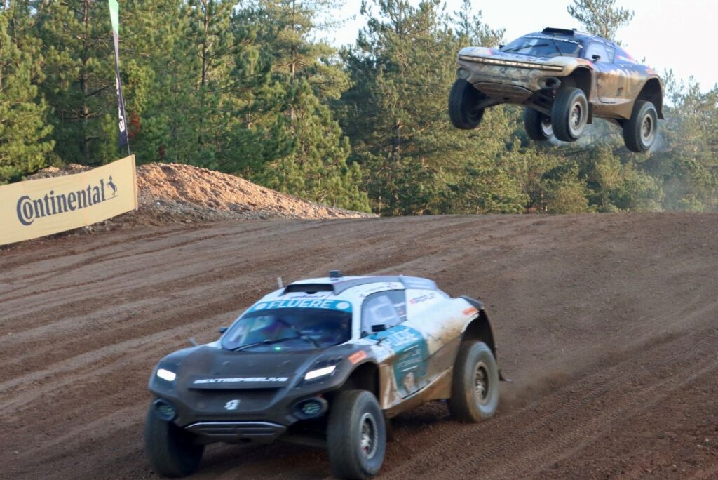 The photo was taken from the side of the racetrack and shows two Extreme E SUVs passing over a jump. The car in front is on the descent while the other follows closely behind in mid-air over the jump. In the background is a row of trees adjacent to the track.