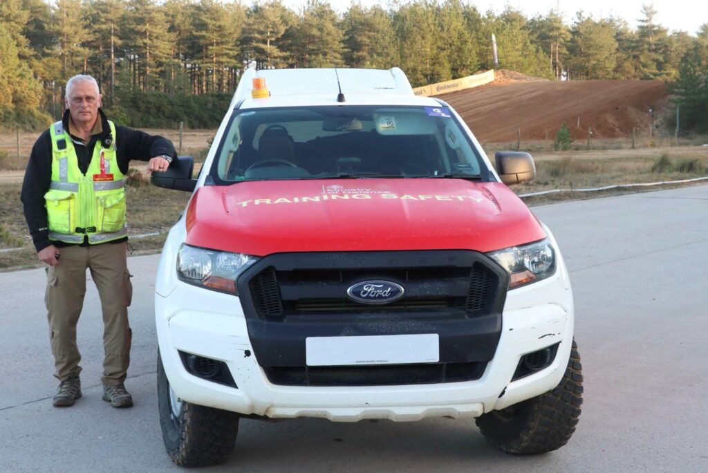 In the centre of the image is a white and red 4x4 vehicle with 'Ministry of Defence Training Safety' written on it. Next to the vehicle is a white male wearing a high-vis jacket. Behind them is a section of the Bovington racetrack, with a row of trees in the background.