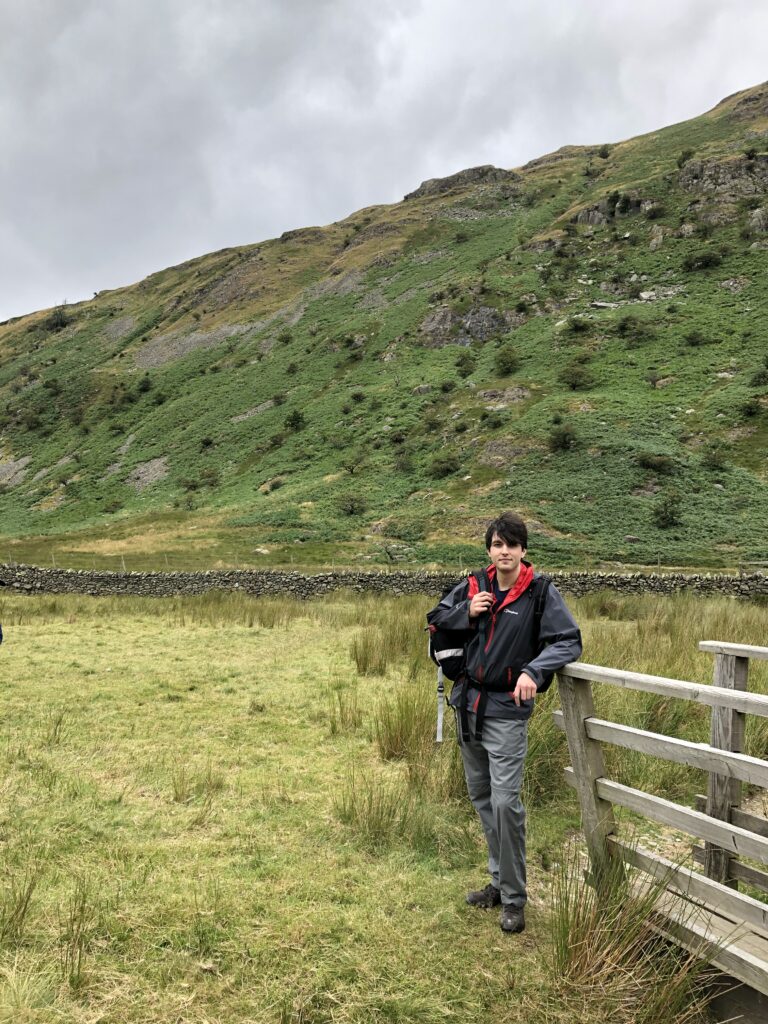 A white man wearing hiking gear and carrying a rucksack stands in an open field next to a wooden fence. In the background there is a dry stone wall with hills behind.