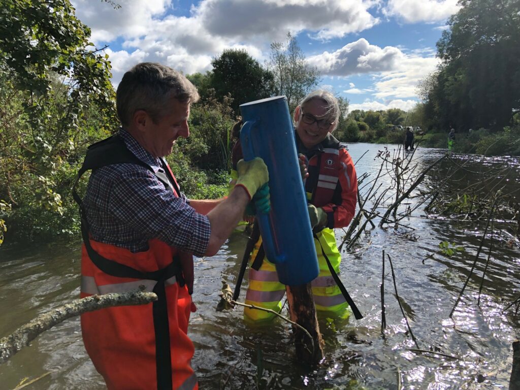 A white male and white woman are stood in waist-high water, wearing waterproof high-vis overalls. They are carrying out maintenance work to tree and branch material in the river.