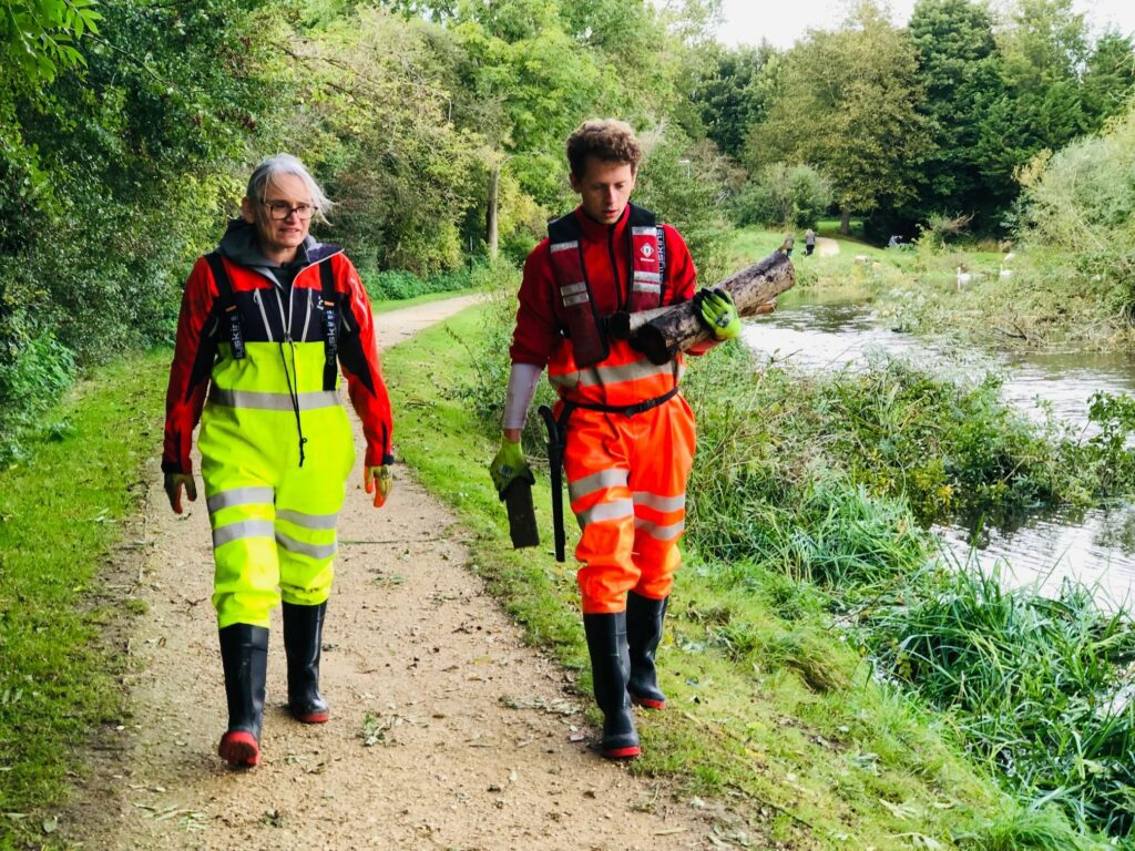 A white female and a white male walk along a riverside path, dressed in high vis clothing and wearing wellies.