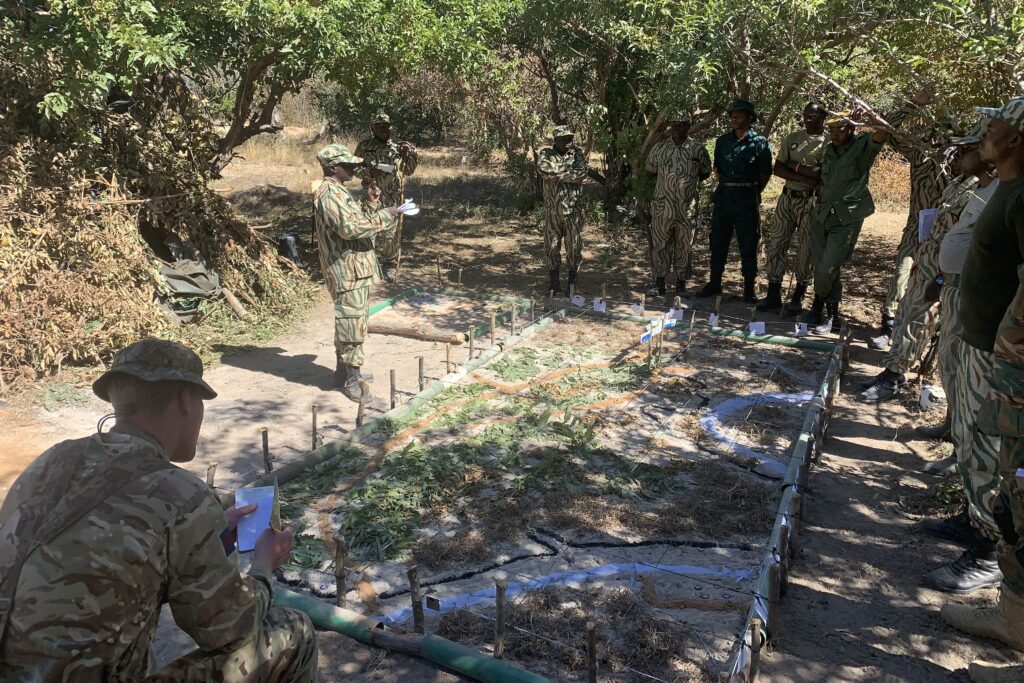 In the bottom left of the image, a white man in military uniform sits with his back to the camera, reading notes. On the ground in front of him is a large map carved into the ground using dirt, sticks and grass. Around the edges of this map are stood a number of black men, also in military fatigues, who are part of the local anti-wildlife poaching ranger force.