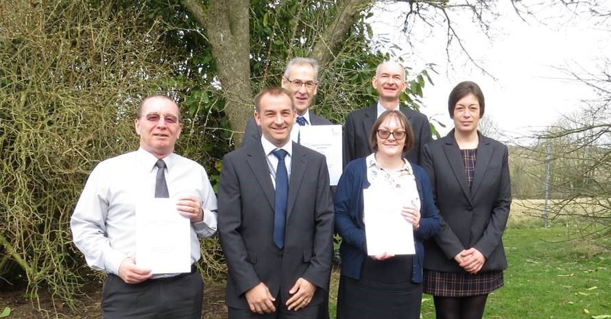 Four white men and two white women are stood facing the camera, presenting award certificates. They are all wearing formal business attire.