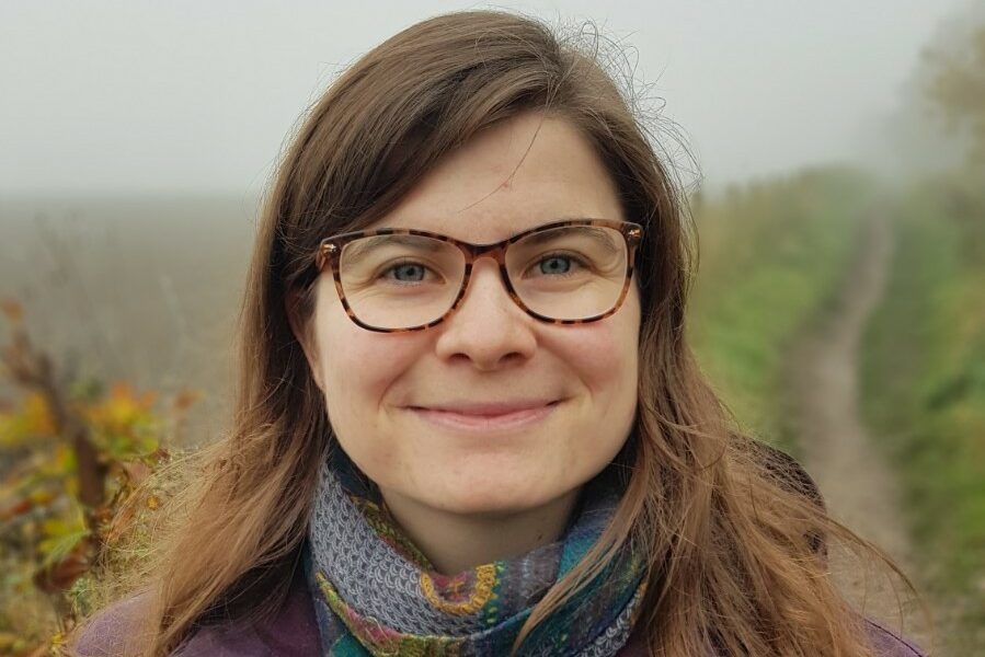 A portrait photo of a white woman with brown hair, who is wearing a scarf. Behind her is a grassy field.