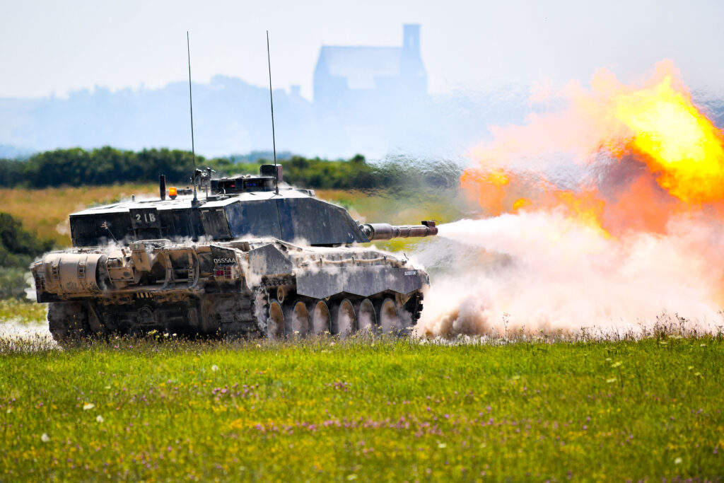 A Challenger 2 tank firing. There is a large explosion and smoke coming from the end of the barrel. The day is sunny.