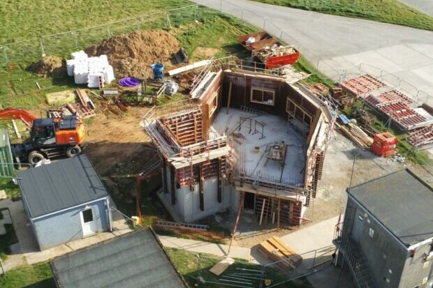 An aerial view of the new tower under construction. The walls are partially complete with the interior being empty. There is a pile of earth in the top of the image.