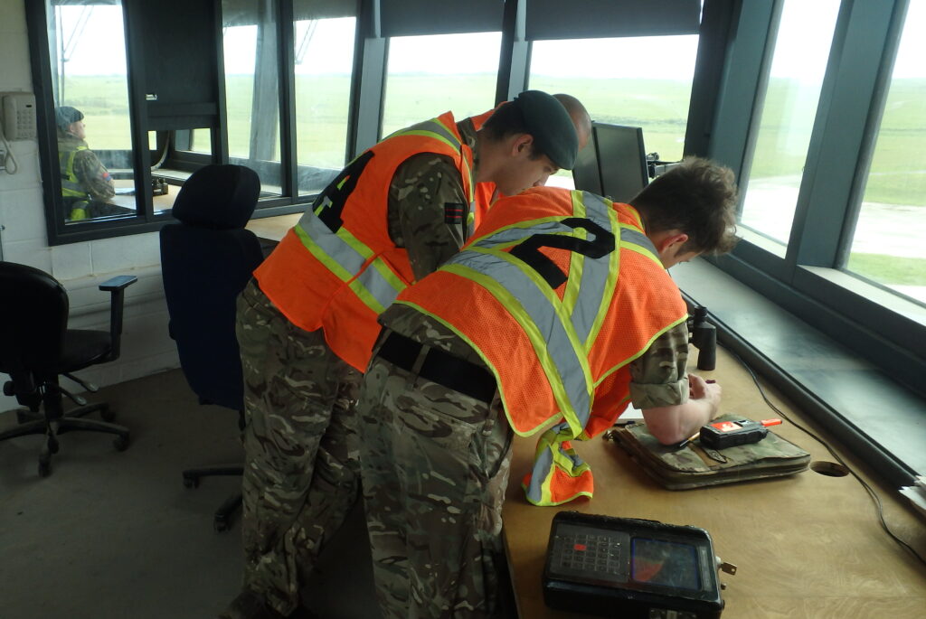 Three soldiers wearing high vis vests with numbers on the back lean over a desk in the control room of the range tower. In front of them are large windows.