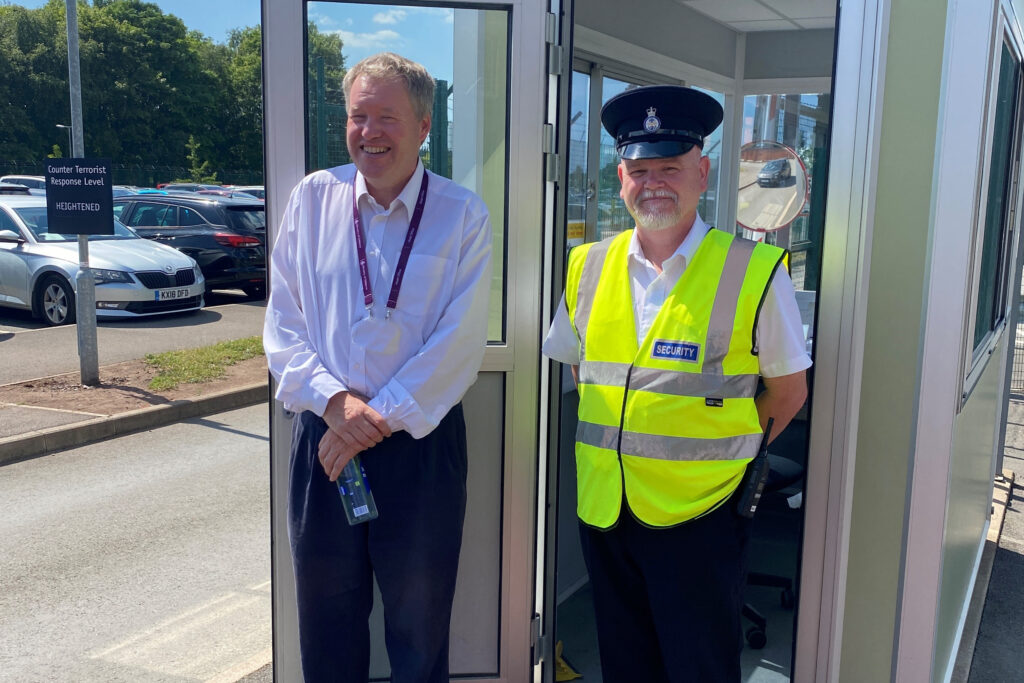 Mike stands next to an MGS security officer, in uniform, outside a security kiosk. 