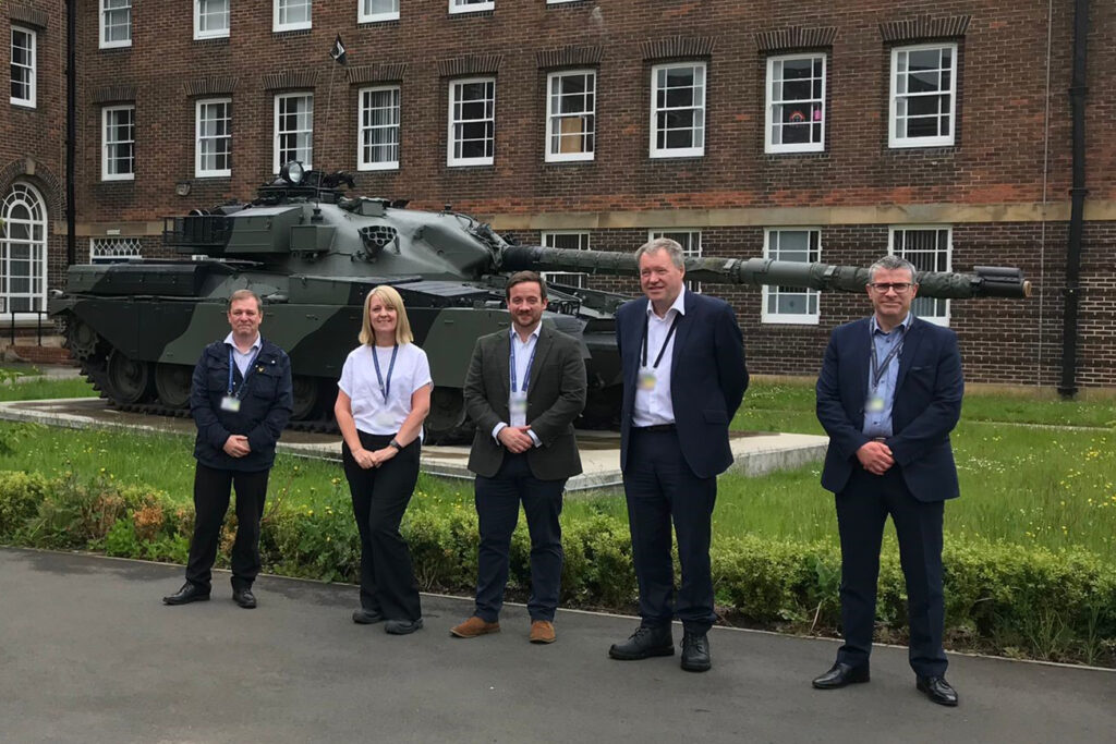 Mike and four other members of DIO staff are lined up in front of a Chieftain tank. Behind them is a grassy area and a large building with many windows.