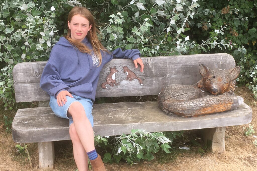 Tristan sitting on a carved wooden bench. Tristan is an 11 year old boy with long hair, wearing a blue hoody and denim shorts.