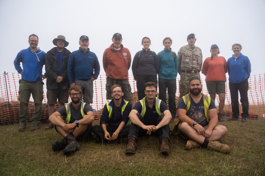 A group of eleven people pose for the camera. Four of them, all wearing t-shirts and high vis vests, sit in front. Seven others stand behind, all casually dressed apart from one who wears military uniform. 