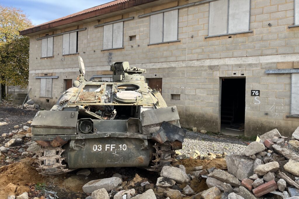 An old and damaged armoured vehicle is on the left of the image, with its tracks up on some rubble which is more prominent to the right of the image. Behind it is a large two storey building made of breeze blocks with shuttered windows.