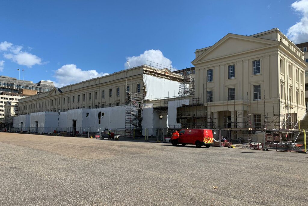 A photo showing the refurbishment work underway, with the bottom half of the section of building shown protected by a cover, with scaffolding around it.