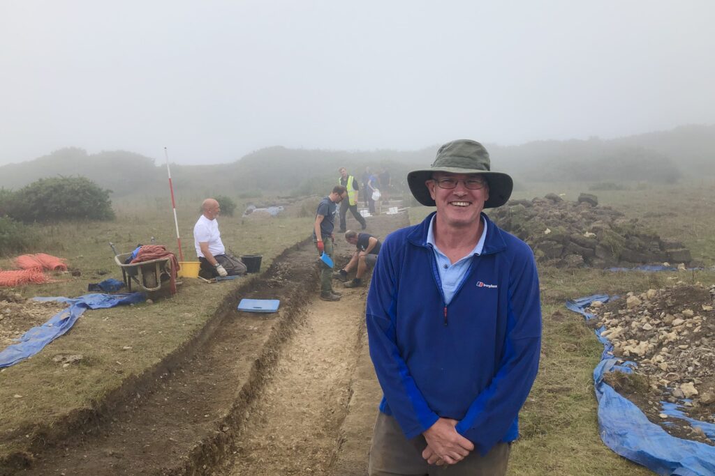 DIO archaeologist Guy Salkeld is pictured in front of a trench under excavation at Flower's Barrow on Lulworth Trainingn Area. He is smiling enthusiastically.