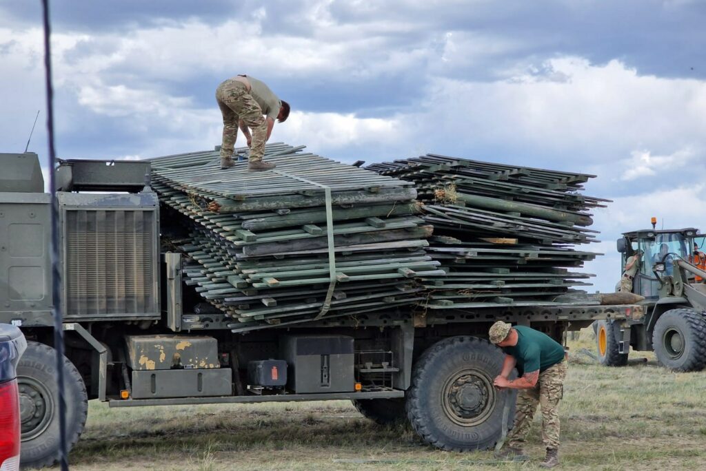 A side-on photo of a large military vehicle loaded with wooden pallets, which are being tied and secured by a pair of men in military uniform.
