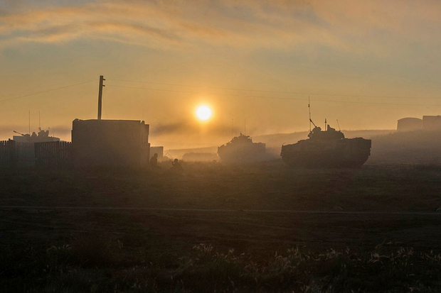 A photo showing a dawn sunrise on the Suffield Military Training Area. Against the dim light can be seen the outlines of several armoured vehicles and some fenced brick buildings.