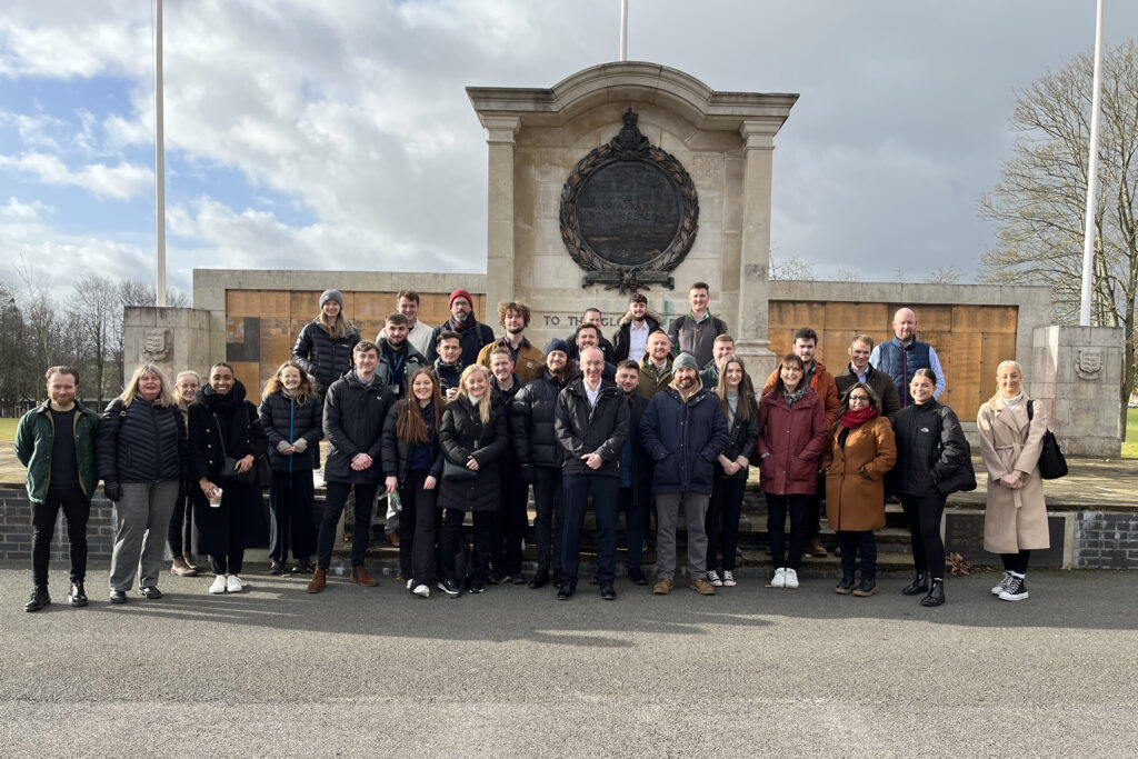 A group of approximately 30 people stand outside, facing the camera, in front of a large memorial with several flagpoles.