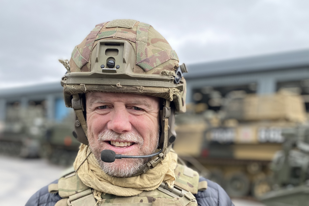 Head and shoulders image of Mike Tindall, a white man with a short beard, smiling at the camera. He wears a military helmet and body armour over a dark jacket.