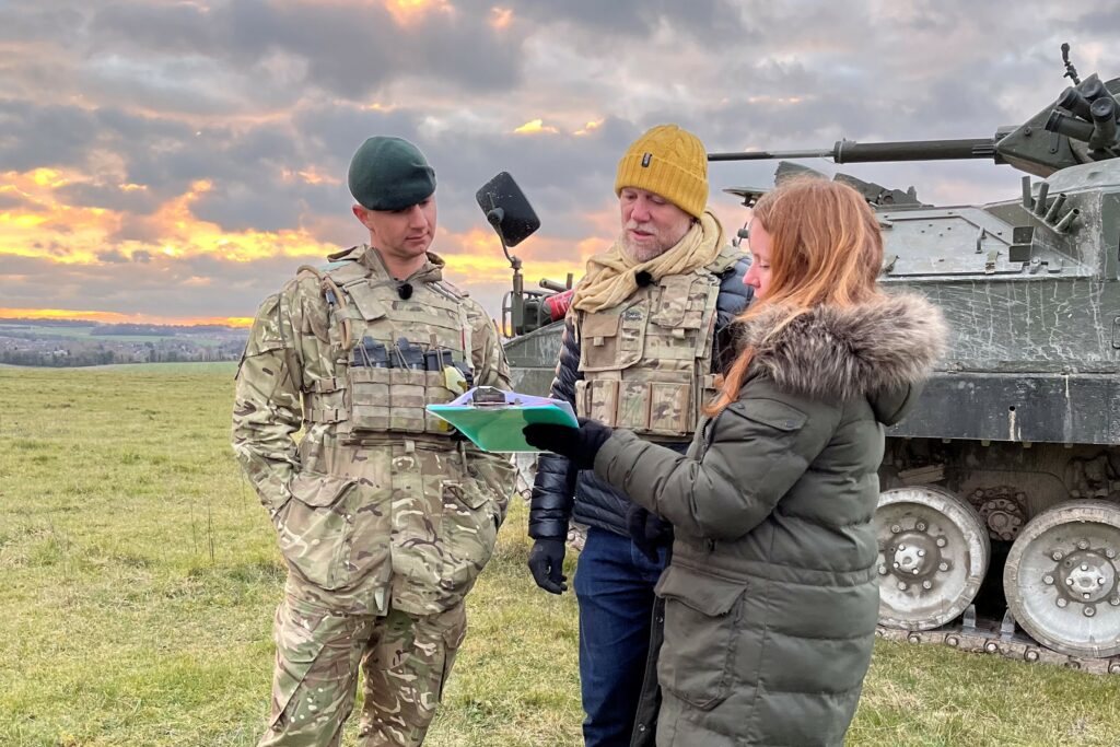 3 people stand in front of a Warrior vehicle looking at a green clipboard. The people and Warrior vehicle are on an area of grassland with a grey sky and orange setting sun in the background