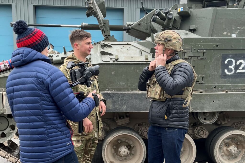 Mike, wearing dark jeans and a jacket with military body armour and helmet, stands next to a Warrior armoured vehicle talking to a soldier. He is being filmed by another man. 