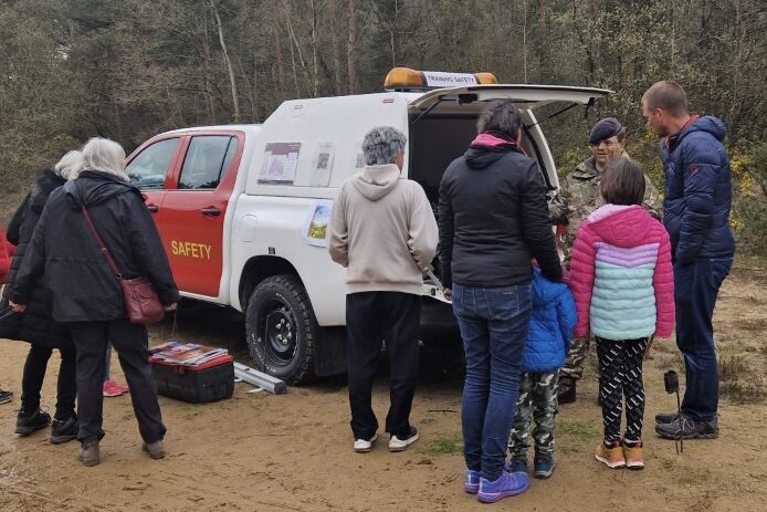 A man in a camouflage military uniform speaking to a group of adults and children. They are stood next to a red and white 4x4 vehicle with 'Training Safety' written on its side.