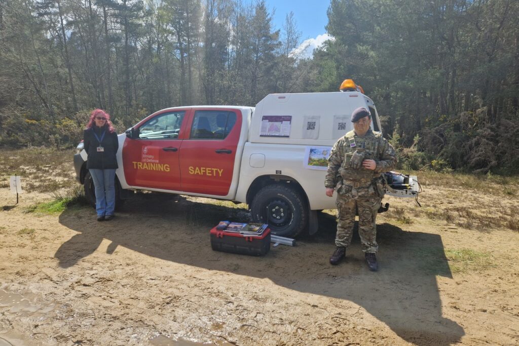 A man in camouflage military uniform and a woman in civilian attire stood in front of a red and white 4x4 vehicle with 'Training Safety' written on its side.