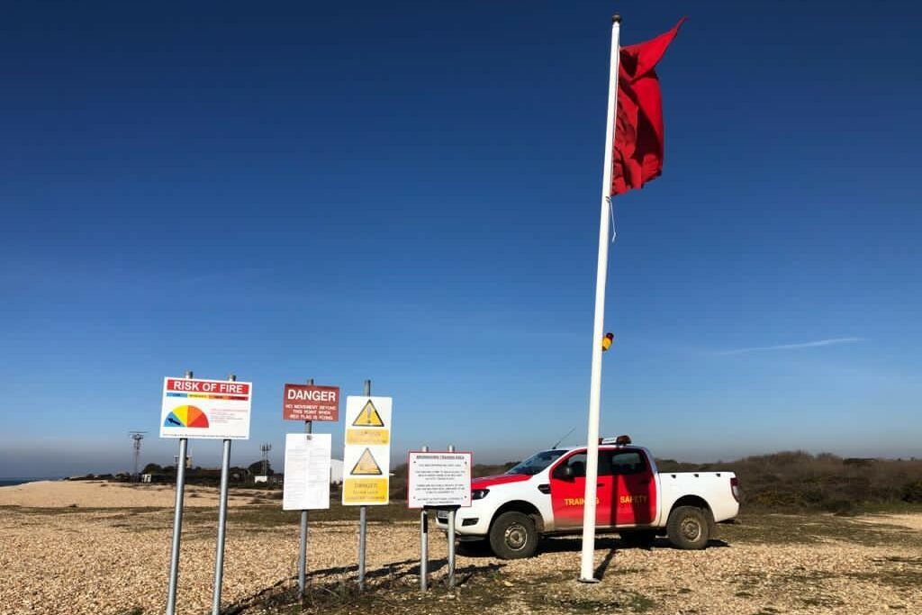 A photo showing a section of beach on a clear, sunny day. On the beach is a flagpole with a red flag flying, several signs with safety information, and a 4x4 vehicle.