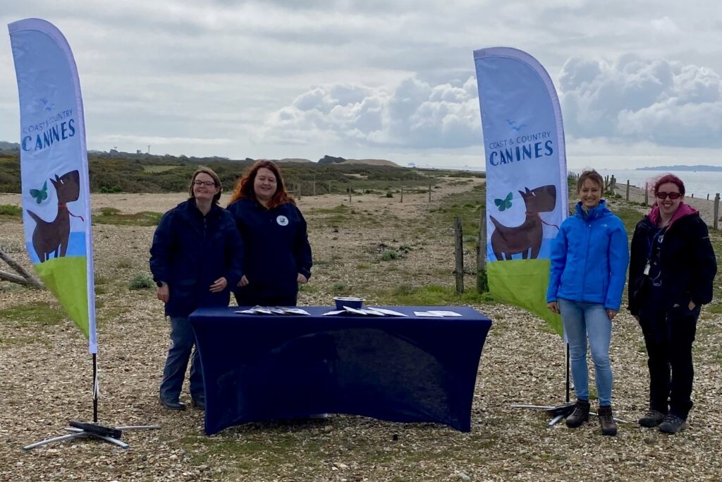 A pair of women in navy blue uniforms stood behind a table on a beach, with flags to their left and right reading 'Coast and Country Canines'. To their left are another pair of women in civilian attire.