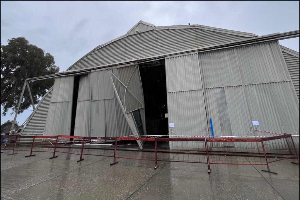 A large, dome-shaped building made of metal, with a large sliding door which is out of place, buckled and damaged.