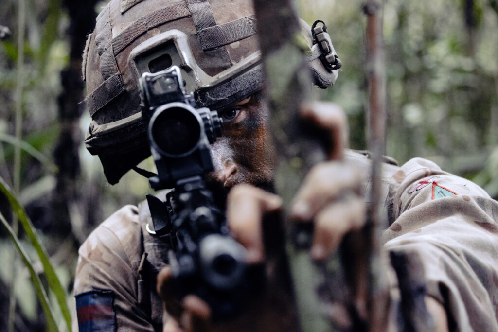 Close up of the head and shoulders of a soldier wearing camouflage uniform and a helmet. He is pointing a rifle directly at the camera and only his left eye can be seen.