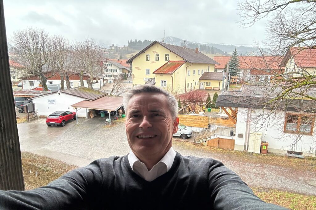 A man in a grey jumper and skirt smiles at the camera in front a road in Germany. The buildings behind him are yellow and white.