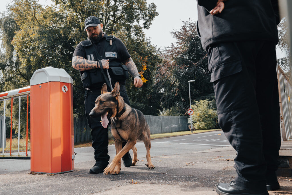 Chris, a bearded white man in a black uniform, walks with Wraith, a dog on a lead, past a red and white vehicle barrier.