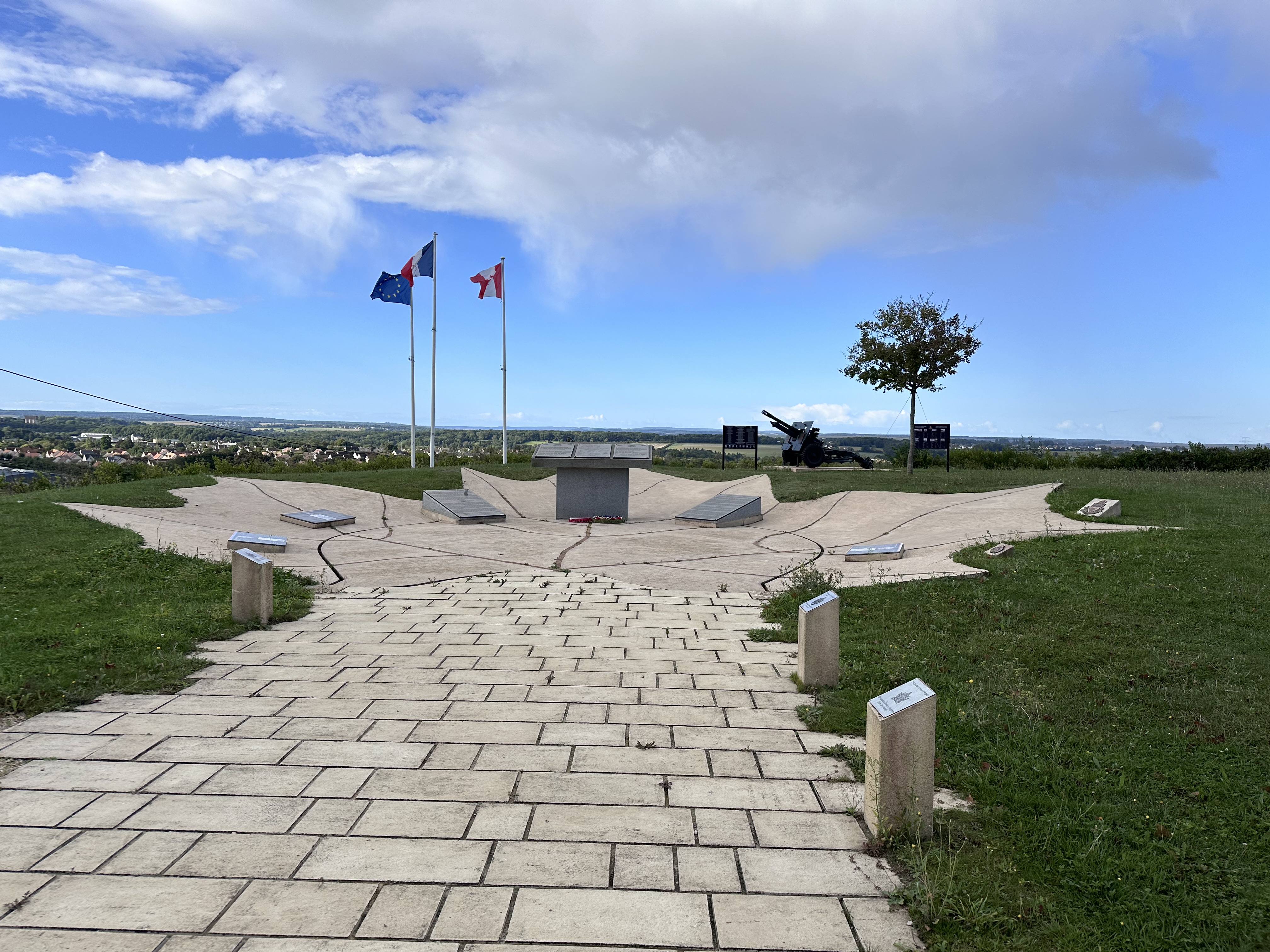 A paved path leading slightly downhill into a paved bowl with the flags of the EU, France and Canada flying over it. 