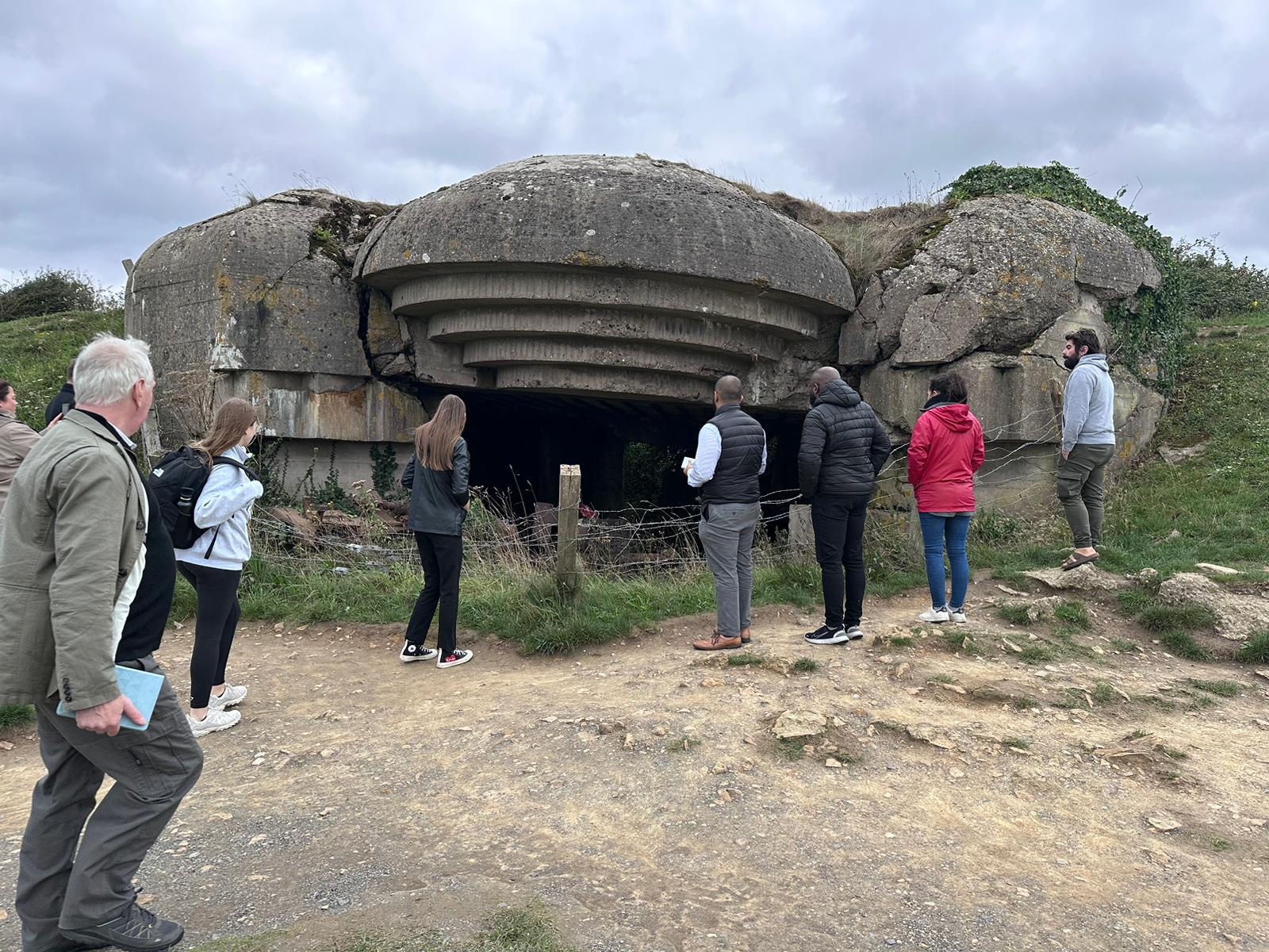 A group of people in casual clothes are gathered around a concrete structure resembling the mouth to a cave.