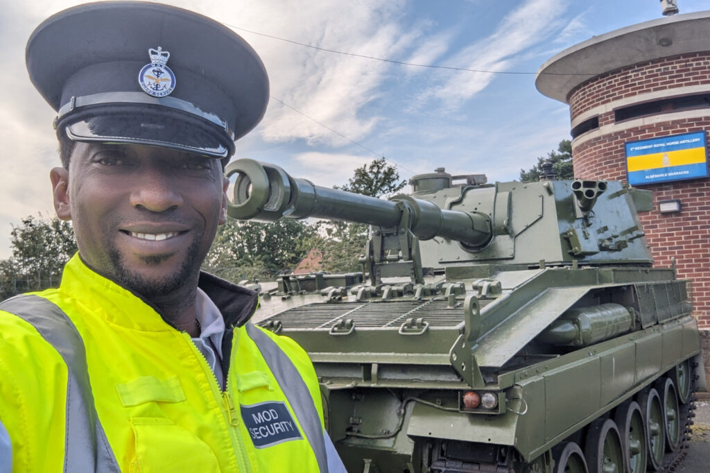 A man wearing a dark peaked hat and a high vis MOD Guard Service jacket smiles at the camera, taking a selfie. Behind him is a tank or armoured vehicle and a round brick building with a sign on it.