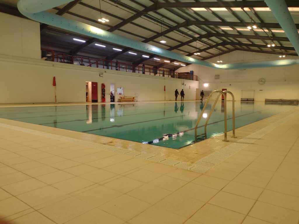 Picture taken from the corner of an indoor swimming pool, showing the tiled edge, the ladder to access the pool, the pool itself and several people in the background.