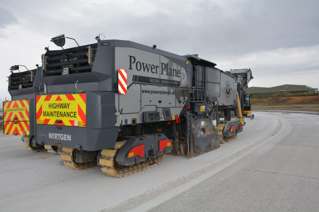 A large vehicle undertaking runway repair work at Mount Pleasant Complex. The vehicle reads 'Power Plane' and has a large 'Highway maintenance' sign on the back. A large green building is just visible in the background. 