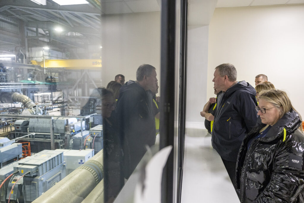 Mike and other visitors look through a window overlooking the interior equipment of the power station, visible to the left of the image.