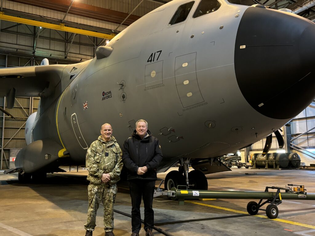 Two men stand in front of a large aircraft in a hangar smiling at the camera. One man wears RAF uniform, the other is in dark civilian clothes.