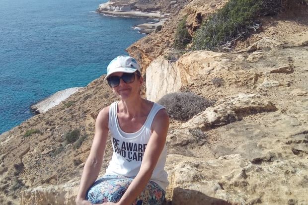 A photo of a woman dressed in a short white top smiling at the camera. She sits in front of a seaside and blue water can be seen behind her.