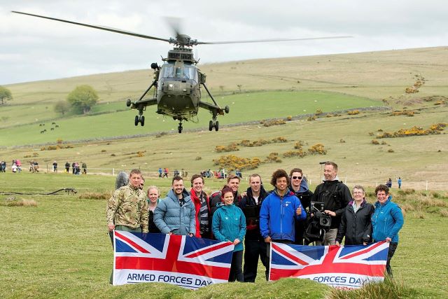 Participants and volunteers at Ten Tors pose with an Armed Forces Day banner as a helicopter flies over them.